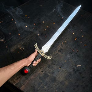 May include: A sword featuring a silver-toned blade with etched line details, a black grip, and a gold-colored crossguard with intricate cutouts. The pommel is circular and contains a prominent, faceted red gemstone set in the center of the base.