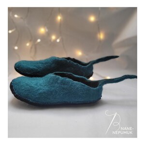 K&ouml;nnte beinhalten: Zwei teal-farbene Filzschuhe mit runder Spitze und einer langen, spitz zulaufenden Verl&auml;ngerung an der Ferse. Die Schuhe sind &uuml;bereinander gestapelt, mit einem schwarzen Rand um die Sohle. Der Hintergrund ist ein weiches Wei&szlig; mit verschwommenen Lichtern.