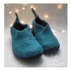 K&ouml;nnte beinhalten: Ein Paar teal-farbene Filzschuhe mit spitzen Spitzen. Die Schuhe haben eine dunkelblaue Sohle und ein weiches, strukturiertes Aussehen. Der Hintergrund zeigt weiche, verschwommene Lichter.