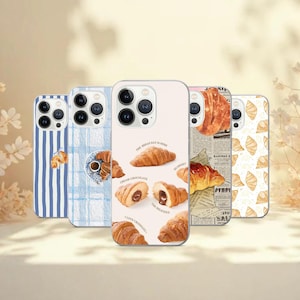 Può includere: Cinque custodie per telefono con disegni di croissant. Una custodia ha strisce verticali blu e bianche, un'altra ha un motivo strutturato blu e bianco con una tazza di caffè e una terza ha uno sfondo crema con illustrazioni di croissant e il testo "The bread day is here".