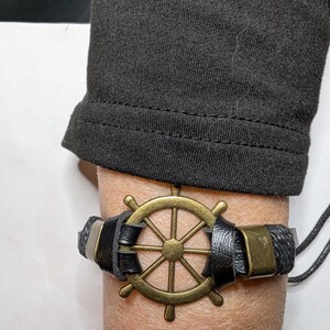 Peut inclure: Un bracelet sur le thème nautique avec un charm en forme de gouvernail de navire de couleur bronze. Le bracelet a un bracelet en cuir noir avec des accents métalliques. Le design évoque une esthétique maritime.