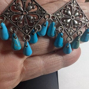 May include: Silver-toned, filigree-style earrings with a diamond-shaped design. Each earring features a central floral motif and is adorned with turquoise teardrop-shaped beads. The earrings are designed for pierced ears.
