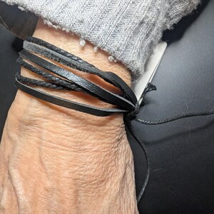 Peut inclure: Bracelet enroulé en cuir noir avec plusieurs brins. Le bracelet est noué avec un cordon noir. Le poignet est couvert par un pull gris.