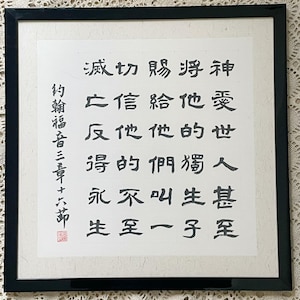 Puede incluir: Arte de caligrafía enmarcado con caracteres de tinta negra sobre fondo blanco. La obra de arte está en un marco negro y presenta texto vertical. El texto incluye las palabras "Juan 3:16". Un sello rojo está en la esquina inferior izquierda.