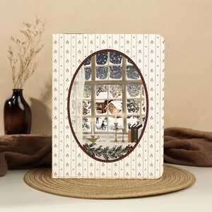May include: A rectangular greeting card featuring a cream-colored background with vertical brown stripes and small floral motifs. An oval cutout in the center reveals a winter scene of a snowy landscape, a house, and figures seen through a window.