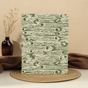 May include: A rectangular notebook featuring a repeating pattern of olive green fish printed on a cream-colored background. The fish are arranged in a dense, overlapping horizontal orientation, covering the entire front cover of the notebook.