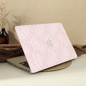 May include: A laptop computer featuring a light pink protective cover with a white floral lace pattern overlay. The cover displays a cutout section revealing the silver Apple logo in the centre of the lid, while the keyboard is visible below.