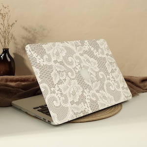 May include: A laptop computer featuring a protective cover with a white lace pattern printed over a beige background. The intricate floral lace design covers the entire outer lid of the device, showcasing a detailed mesh and petal motif.