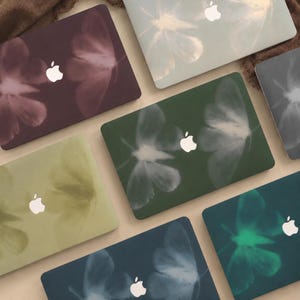 Può includere: Una collezione di cover per laptop con un design ad ali di farfalla in vari colori, tra cui prugna intenso, verde salvia, verde bosco, oliva, ottanio e grigio antracite, ciascuna con un logo Apple bianco centrato sulla superficie.