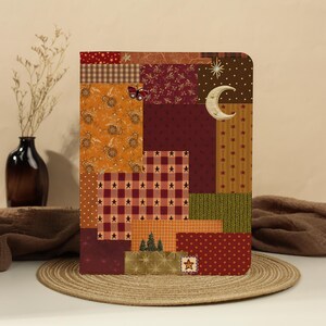 Peut inclure: Un carnet rectangulaire avec une couverture style patchwork présentant divers motifs dont des carreaux, des étoiles, un croissant de lune, un papillon et des sapins dans des tons bordeaux, orange, marron et doré. Les coins sont arrondis.