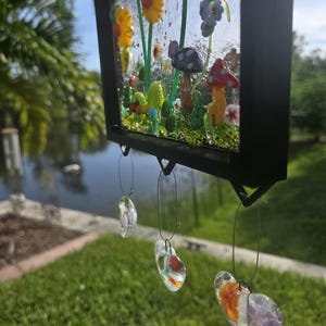 May include: A rectangular stained glass suncatcher in a black frame featuring a garden scene with yellow sunflowers, red and purple mushrooms, and green foliage. Three clear glass charms with orange and purple accents hang from the bottom.
