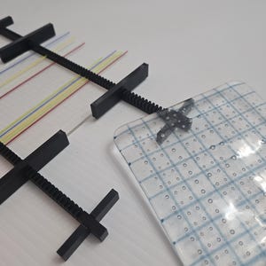 May include: Black plastic weaving loom with colorful threads and a clear, square, grid-patterned piece. The loom has a series of crossbars and a threaded rod. The clear piece has a blue grid pattern.