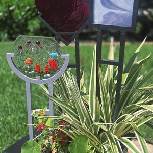May include: A decorative garden art piece featuring a heart-shaped red glass element, a rectangular panel, and a glass aquarium-like structure with colorful aquatic-themed decorations. The art is set in a planter with greenery.