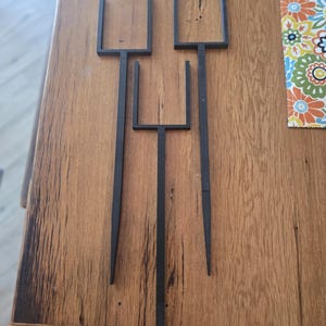 May include: Three black metal decorative garden stakes with a unique, minimalist design. Each stake has a long, pointed base and a rectangular top with two vertical bars. The stakes are arranged on a wooden surface.
