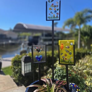 May include: Three decorative garden stakes with colorful glass panels featuring floral and butterfly designs. The stakes are set in a blue ceramic pot with greenery. The panels are framed in black metal and stand against a bright blue sky.