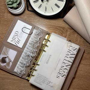 May include: A taupe leather planner with gold accents, open to reveal a savings challenges insert. The planner includes a lace divider, a card that says "Mindful", and a small photo. A black clock and a small succulent are in the background.