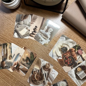 May include: A collection of aesthetic art cards featuring various lifestyle images. The cards showcase scenes with books, food, and decor in neutral tones. A black clock and a small succulent in a white pot are also visible.