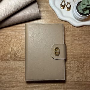 May include: A beige leather-like planner with a gold-colored clasp is centered on a wooden surface. Several other planners in shades of beige and white are stacked to the left. A white tray with jewelry and a small succulent plant is to the right.