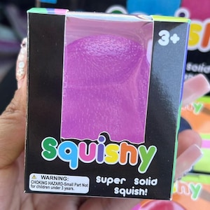 May include: A purple squishy toy in a box. The box has the word "squishy" in colourful letters. The box also has a warning label and the words "super solid squish!"