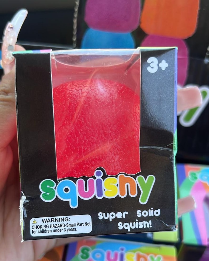 May include: A red squishy toy in a black box with the word "squishy" in colourful letters. The box also has the text "super solid squish!" and a warning about choking hazards for children under 3 years old.