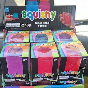 May include: A display of "Squishy" toys in boxes. The boxes are black with the word "Squishy" in green and the text "super solid squish!". The toys are in various colours, including pink, red, and blue. The display also has a cardboard cutout with two squishy shapes.