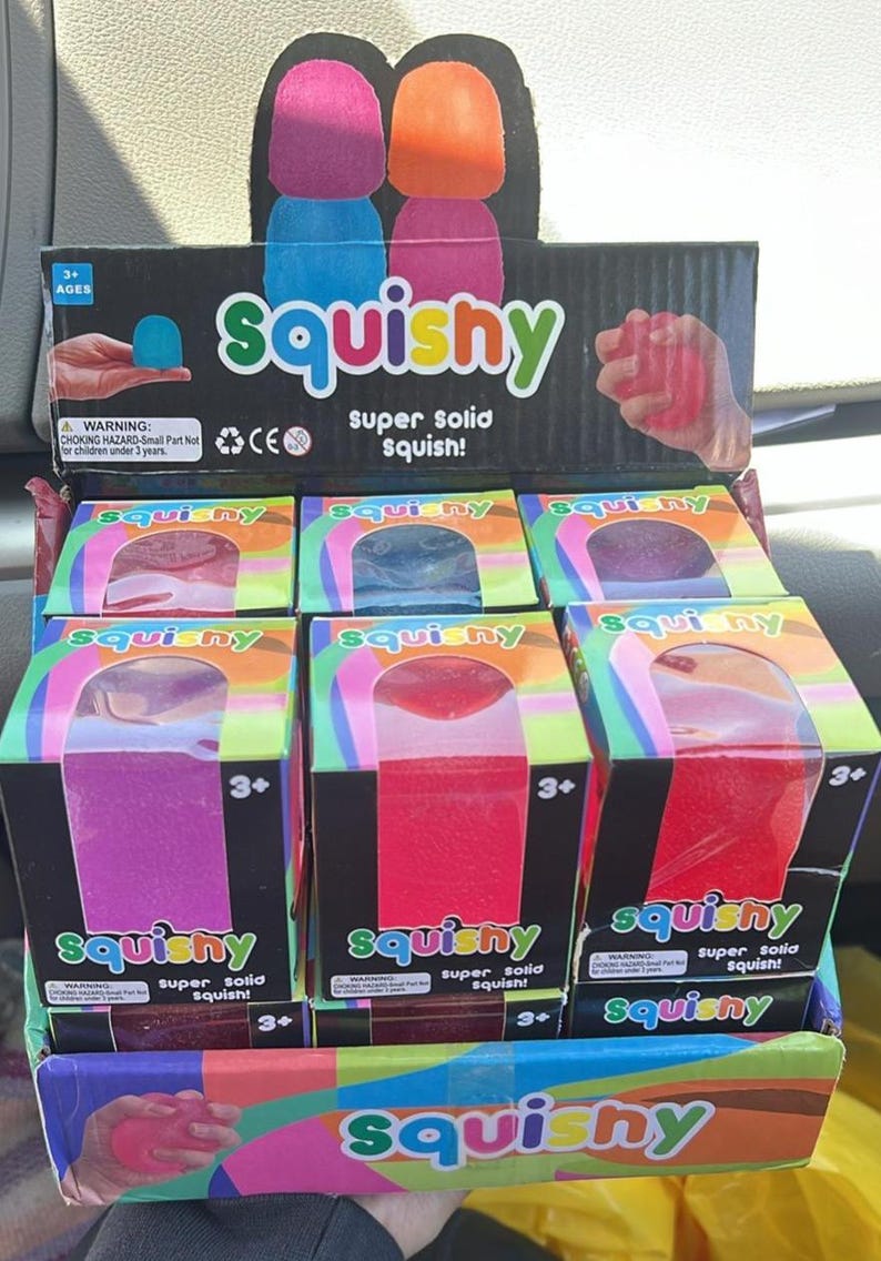 May include: A display of "Squishy" toys in boxes. The boxes are black with the word "Squishy" in green and the text "super solid squish!". The toys are in various colours, including pink, red, and blue. The display also has a cardboard cutout with two squishy shapes.