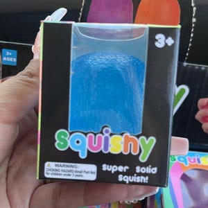 May include: A blue squishy toy in a black box. The box features the word "squishy" in green and orange, and the text "super solid squish!". The box also has a warning for children under 3 years old.