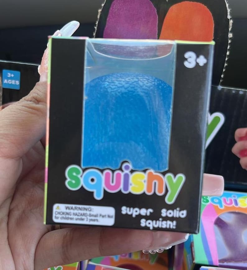 May include: A blue squishy toy in a black box. The box features the word "squishy" in green and orange, and the text "super solid squish!". The box also has a warning for children under 3 years old.