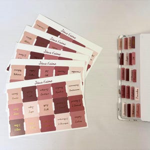 Peut inclure: Un ensemble d'onglets adhésifs pour Bible dans des tons de rose, mauve et bordeaux, portant le texte 'Jésus t'aime' et divers noms de livres. Les onglets sont disposés sur des feuilles et également appliqués sur le côté d'une page lignée.