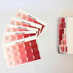 Peut inclure: Un ensemble d'onglets adhésifs pour Bible disposés en éventail, présentant un dégradé de tons rouges et roses avec le texte JÉSUS T'AIME imprimé en haut. Un livre est visible sur la droite avec plusieurs onglets appliqués sur les bords.
