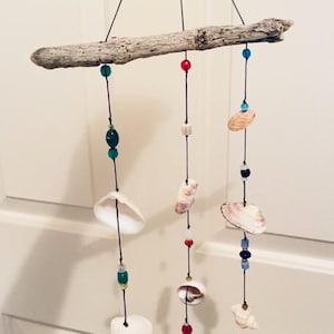 May include: A driftwood mobile with seashells and colourful beads. The natural wood branch suspends strands of black cord, each adorned with shells, green, blue, red, and white beads. A coastal-themed decorative item.