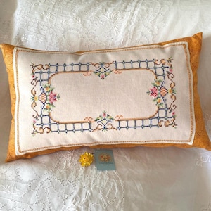 May include: A rectangular throw cushion featuring a central cream-coloured panel with a cross-stitched border of blue grid lines and floral motifs in pink, yellow, and green. The cushion has mustard-yellow side panels and a small blue product tag.
