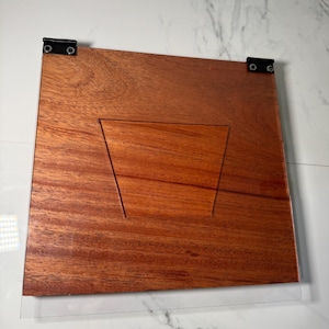 May include: A wooden display stand with a clear acrylic front. The stand is made of reddish-brown wood and features a trapezoid-shaped cutout. Black hinges secure the wood to the acrylic. The stand is designed to hold and display items.