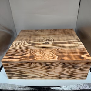 May include: A square wooden box with a dark, scorched wood grain finish. The lid features a circular logo with a tree illustration and the text ARRIOLA centered on the top surface. The box has a flat top and straight sides with visible wood texture.