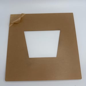 May include: A square, brown acrylic sheet with a trapezoid-shaped cutout in the center. A protective film is partially peeled back from the upper left corner. The background is plain white.