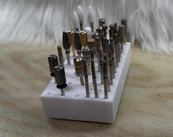 May include: A white rectangular nail drill bit holder filled with various metal drill bits. The bits have different shapes and sizes, including cylindrical, cone, and ball shapes. Some bits have gold or silver accents.