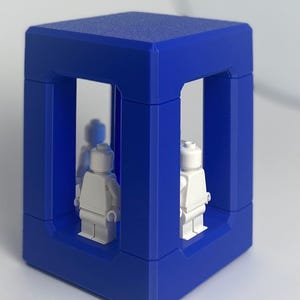 May include: A cobalt blue cube-shaped display case with open sides, showcasing two white toy figures. The case has a textured surface and a modern design. The figures are in a standing position.