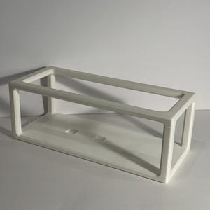 Può includere: Un espositore rettangolare bianco a telaio aperto. Lo stand ha una struttura a forma di scatola con una base solida e una parte superiore aperta. Il telaio è realizzato in un materiale liscio e opaco. Lo stand è progettato per contenere ed esporre oggetti.