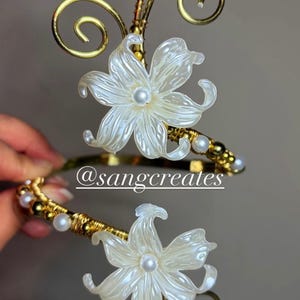May include: Gold-toned floral hair accessory featuring a white flower with a pearl center. The design includes gold wire detailing and small pearls. The accessory has a delicate, elegant appearance, suitable for special occasions.