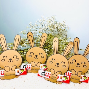 May include: Four brown paper bunny cutouts with smiling faces and long ears, each holding a chocolate bar. The bars have red and white wrappers with text. White flowers and a light blue background complete the Easter-themed arrangement.