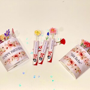 Op de afbeelding: Een verjaardagsarrangement met papieren bloemboeketten en drie Kinder Surprise-eieren met bloemaccenten. De boeketten zijn versierd met bloemmotieven en de teksten "HAPPY BIRTHAY" en "Mit liebe".