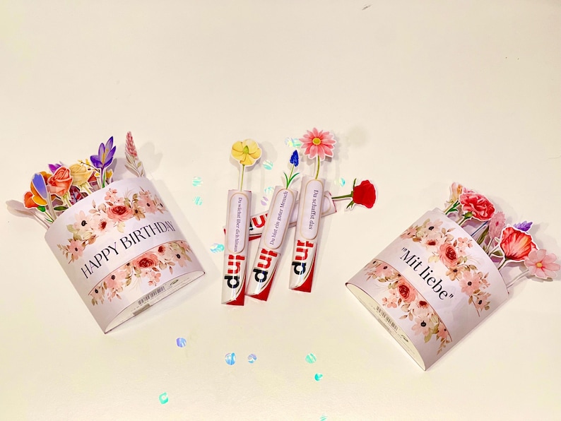 Op de afbeelding: Een verjaardagsarrangement met papieren bloemboeketten en drie Kinder Surprise-eieren met bloemaccenten. De boeketten zijn versierd met bloemmotieven en de teksten "HAPPY BIRTHAY" en "Mit liebe".