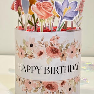 Op de afbeelding: Een witte, cilindrische geschenkdoos met een bloemenontwerp en de tekst "HAPPY BIRTHAY". De doos is versierd met papieren bloemen in verschillende kleuren, waaronder roze, geel en paars. Het thema is feestelijk.