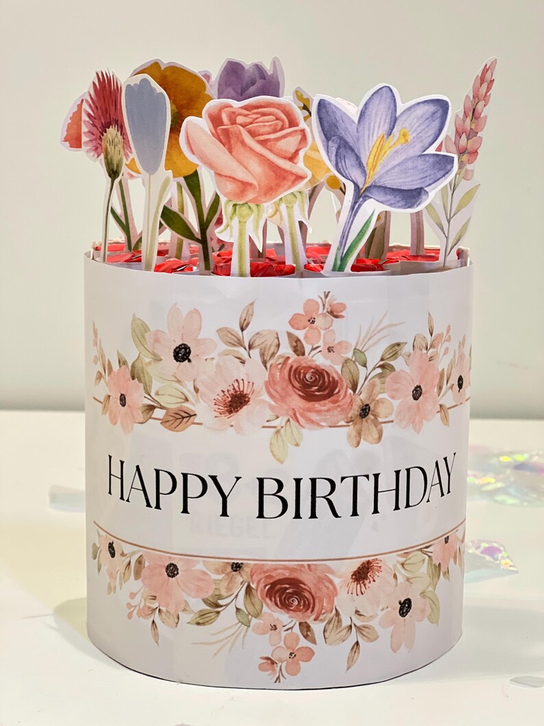 Op de afbeelding: Een witte, cilindrische geschenkdoos met een bloemenontwerp en de tekst "HAPPY BIRTHAY". De doos is versierd met papieren bloemen in verschillende kleuren, waaronder roze, geel en paars. Het thema is feestelijk.