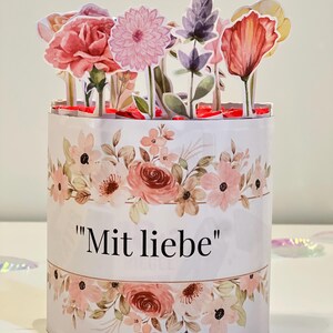 Op de afbeelding: Een cilindrische bloemen geschenkdoos met een pop-up bloemstuk. De doos is wit met een aquarel bloemenpatroon in roze en perziktinten. De tekst "Mit liebe" staat op de voorkant. De pop-up bloemen omvatten anjers, dahlia's en andere bloemen.