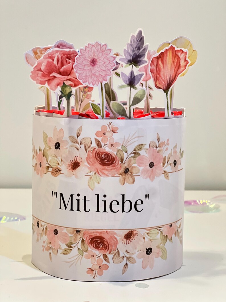 Op de afbeelding: Een cilindrische bloemen geschenkdoos met een pop-up bloemstuk. De doos is wit met een aquarel bloemenpatroon in roze en perziktinten. De tekst "Mit liebe" staat op de voorkant. De pop-up bloemen omvatten anjers, dahlia's en andere bloemen.