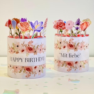 Op de afbeelding: Twee cilindrische bloemrijke geschenkdozen. Op &eacute;&eacute;n staat "HAPPY BIRTHDAY" en op de andere "Mit liebe". Beide hebben een bloemenontwerp en zijn bekroond met papieren bloemen in verschillende kleuren. De dozen staan op een wit oppervlak met confetti.