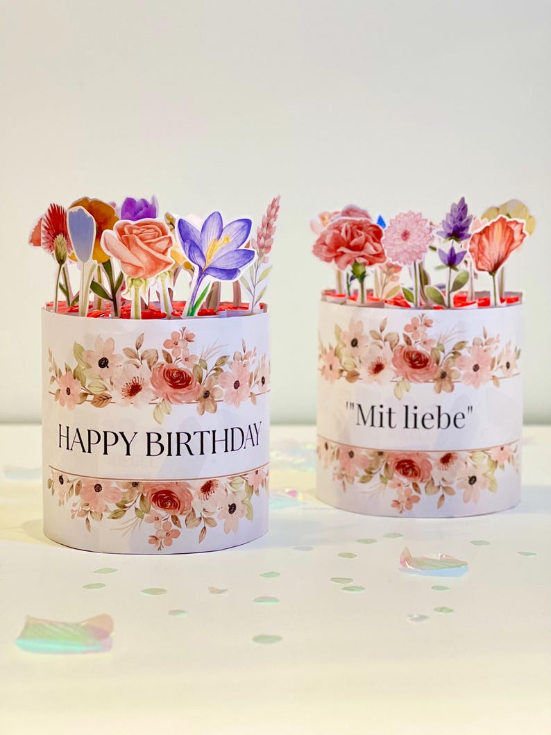 Op de afbeelding: Twee cilindrische bloemrijke geschenkdozen. Op &eacute;&eacute;n staat "HAPPY BIRTHDAY" en op de andere "Mit liebe". Beide hebben een bloemenontwerp en zijn bekroond met papieren bloemen in verschillende kleuren. De dozen staan op een wit oppervlak met confetti.