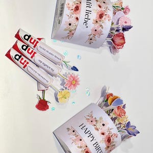 Op de afbeelding: Drie snoeprepen met het woord "inp" erop gedrukt, naast twee met bloemen versierde papieren bakjes. Op &eacute;&eacute;n bakje staat "Mit liebe" en op de andere "HAPPY BIRTHDAY". Kleurrijke papieren bloemen komen uit de bakjes.