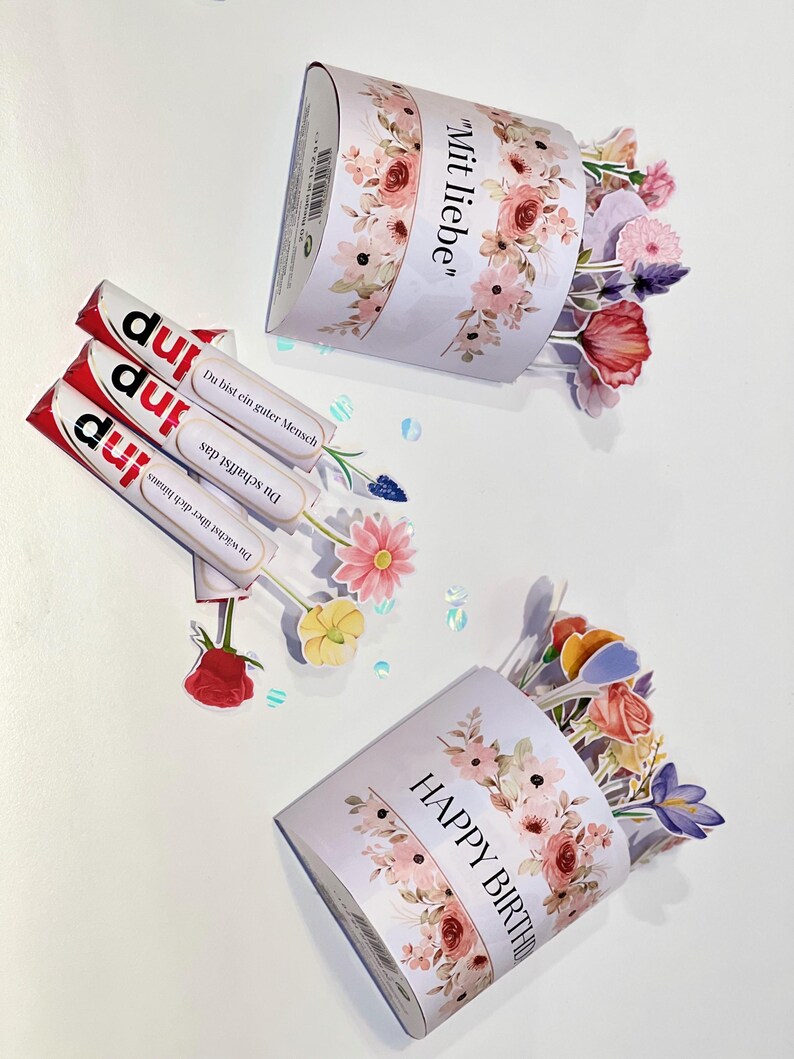 Op de afbeelding: Drie snoeprepen met het woord "inp" erop gedrukt, naast twee met bloemen versierde papieren bakjes. Op &eacute;&eacute;n bakje staat "Mit liebe" en op de andere "HAPPY BIRTHDAY". Kleurrijke papieren bloemen komen uit de bakjes.